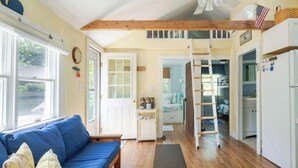 Una habitación acogedora con un sofá azul, piso de madera, una escalera que lleva a un altillo y un refrigerador.
