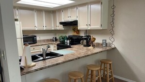 A kitchen with white cabinets, a black stove, a microwave, and a sink. There are three wooden stools at the counter. - Schweitzer Mountain Condo (Sandpoint)