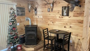 A cozy cabin interior with a wood-burning stove, a dining table with chairs, a decorated Christmas tree, and mounted animal skulls on the walls.