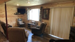 A cozy cabin interior with a wooden floor, a fireplace, a television mounted on the wall, and a window with curtains.