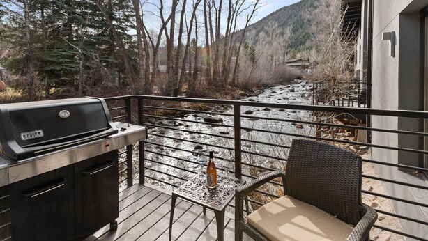 Una terraza con una parrilla, una botella y una silla, con vistas a un río y colinas boscosas.