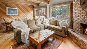 Una sala de estar acogedora con techo de madera, chimenea de ladrillo y un ventanal con vistas a un bosque.