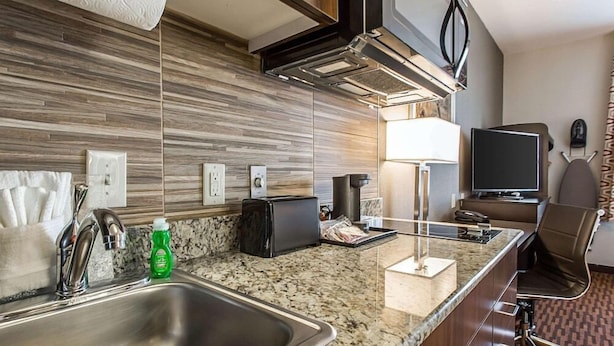 A modern kitchen with a granite countertop, stainless steel sink, and a microwave. There is a chair and a television in the corner. - Suburban Studios Midland I-20 (Midland)