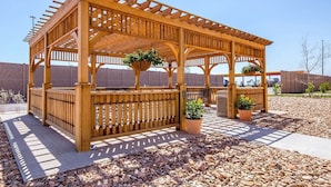 A wooden gazebo with a lattice roof, surrounded by a brick wall, and featuring outdoor seating and planters. - Suburban Studios Midland I-20 (Midland)