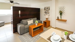 A modern living room with a grey sofa, wooden side tables, a patterned rug, and a dining area with white plates and a vase of flowers. - Zimmer Suites (Aguascalientes)