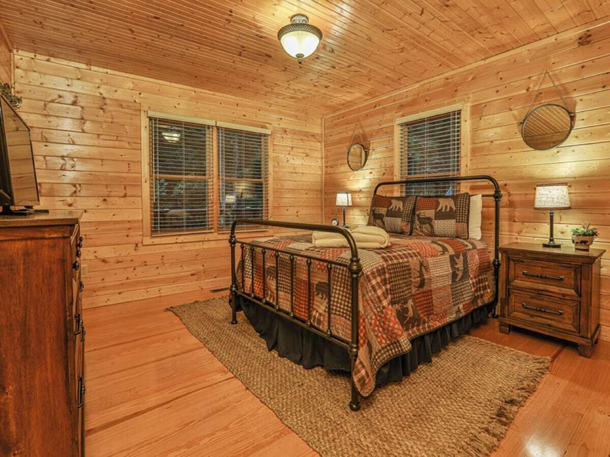 A log cabin bedroom with a metal bed frame, a wooden dresser, and a nightstand with a lamp.