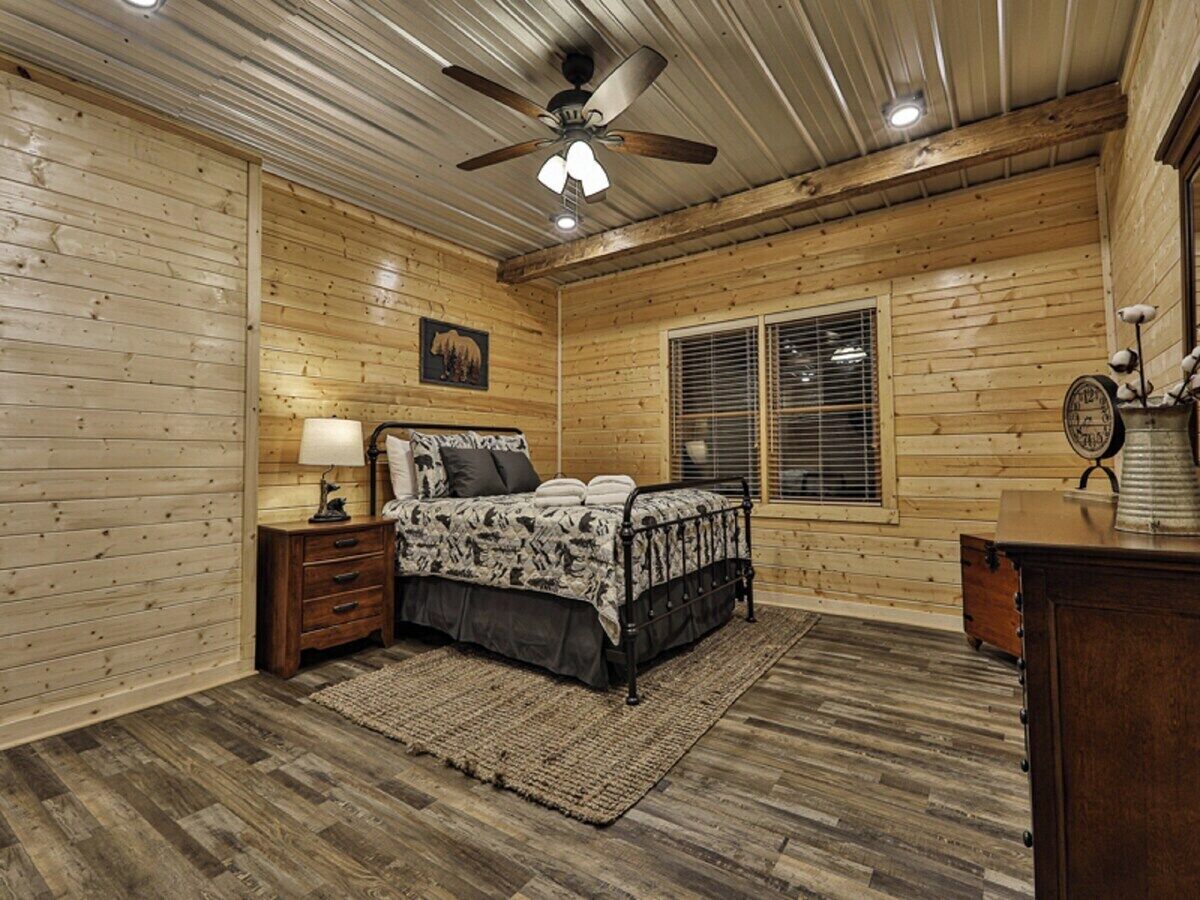 Un dormitorio con cama, paredes de madera, ventilador de techo, ventana con persianas y mesita de noche con lámpara.