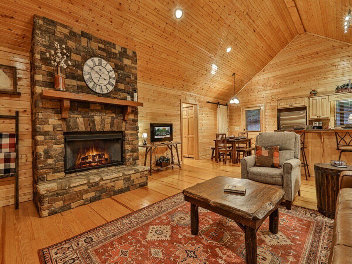 A cozy log cabin interior with a stone fireplace, wooden walls, a dining area, and a living room furnished with a coffee table and armchairs.