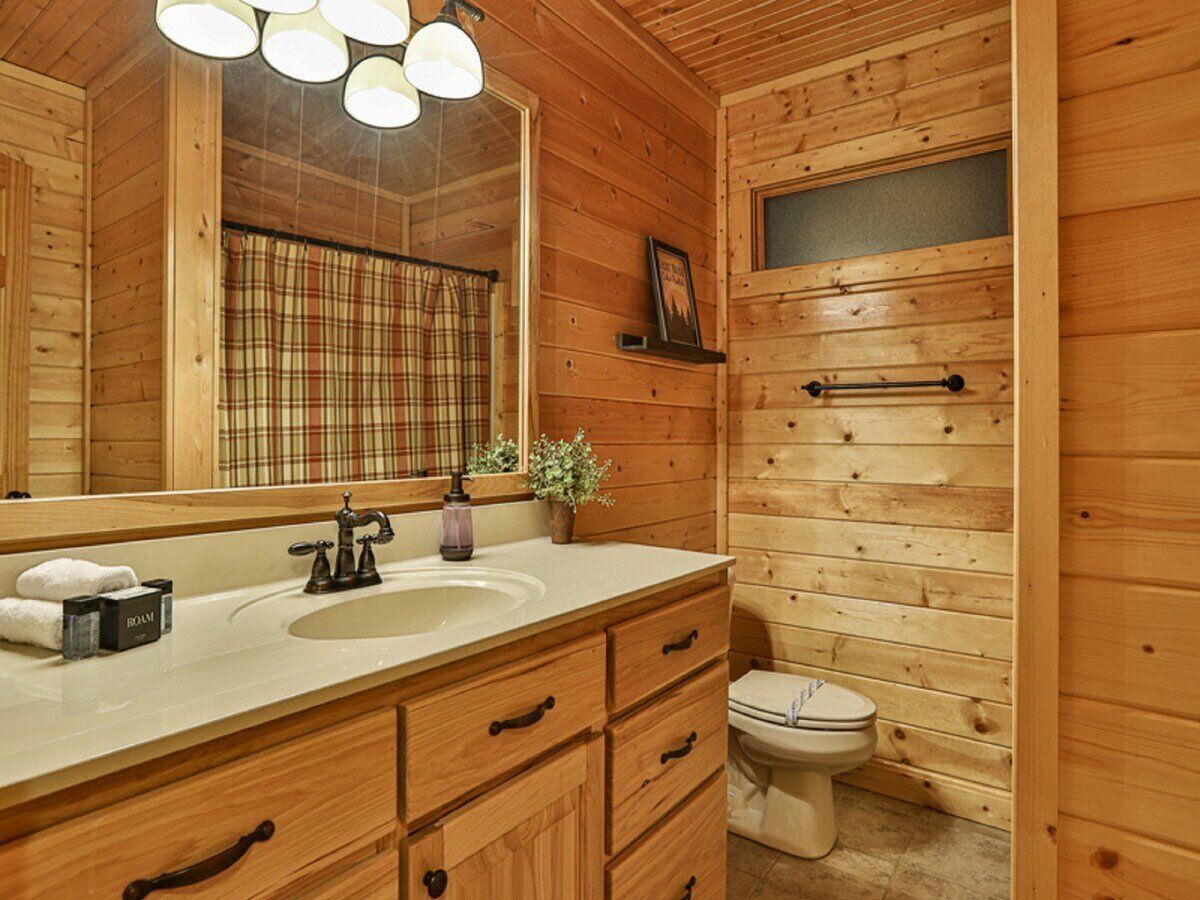 A wooden cabin bathroom with a large mirror, a sink, a toilet, and a plaid curtain.