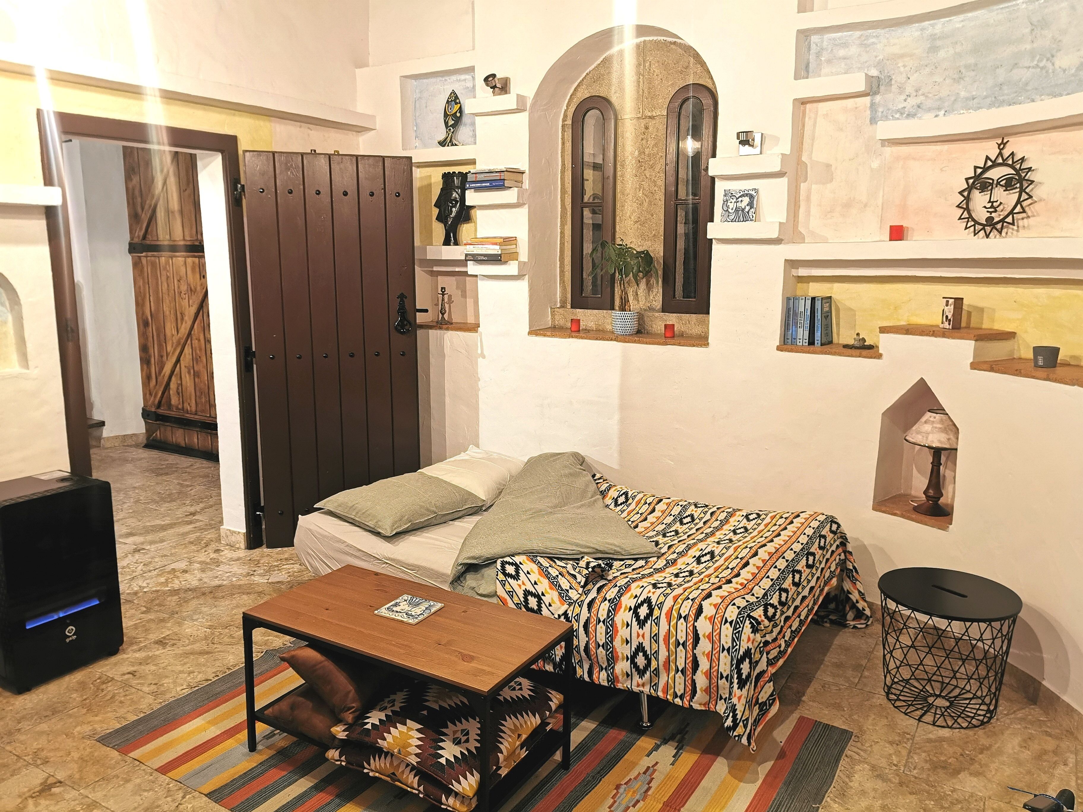 Una habitación pequeña y acogedora con una cama, una mesa de madera, un estante con libros, una lámpara y un adorno en la pared.