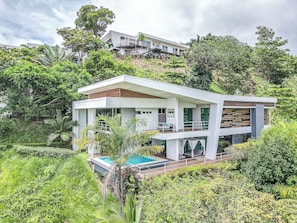 A modern two-story house with a pool, surrounded by lush greenery and hills.