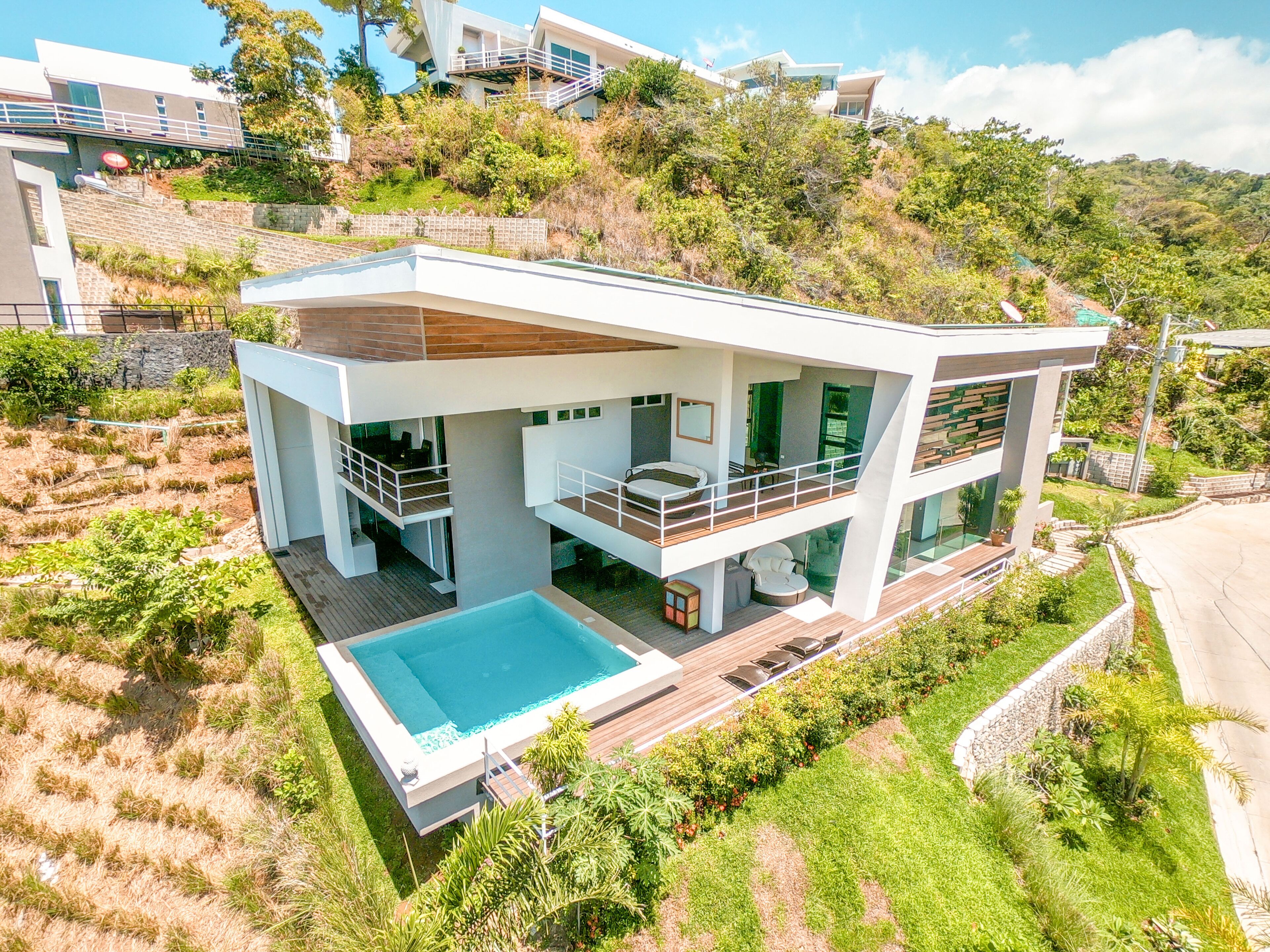 A modern two-story house with a swimming pool, multiple balconies, and a lush garden.