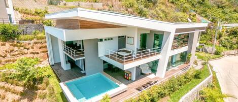 A modern two-story house with a swimming pool, multiple balconies, and a lush garden.