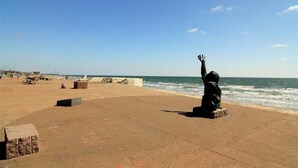 En strand med en staty av en person som lyfter handen, en klar himmel och havet i bakgrunden.