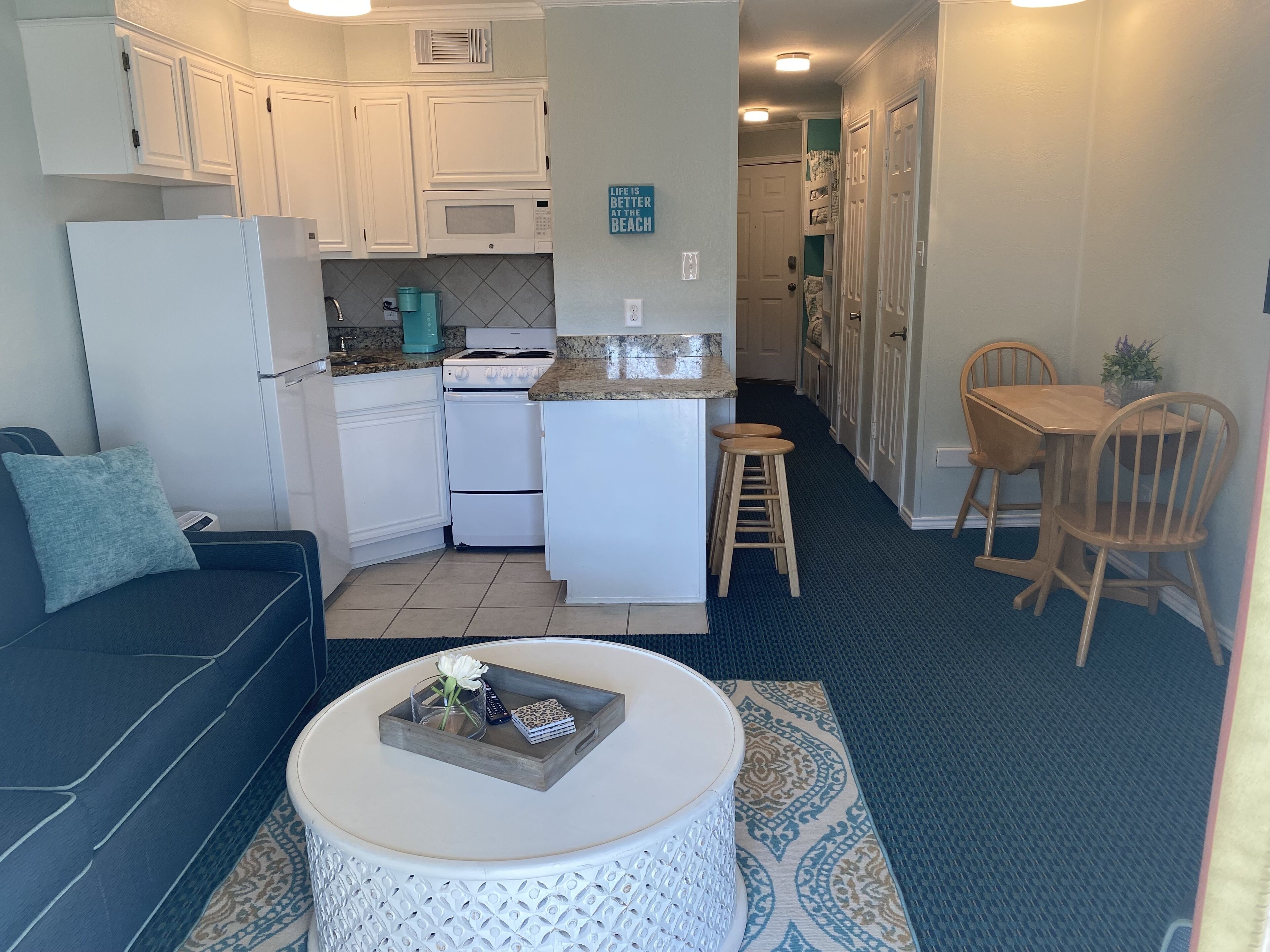 A compact kitchen with white cabinets, a microwave, stove, and a coffee maker. There is a dining area with a table and chairs, and a small seating area with a couch and a coffee table.