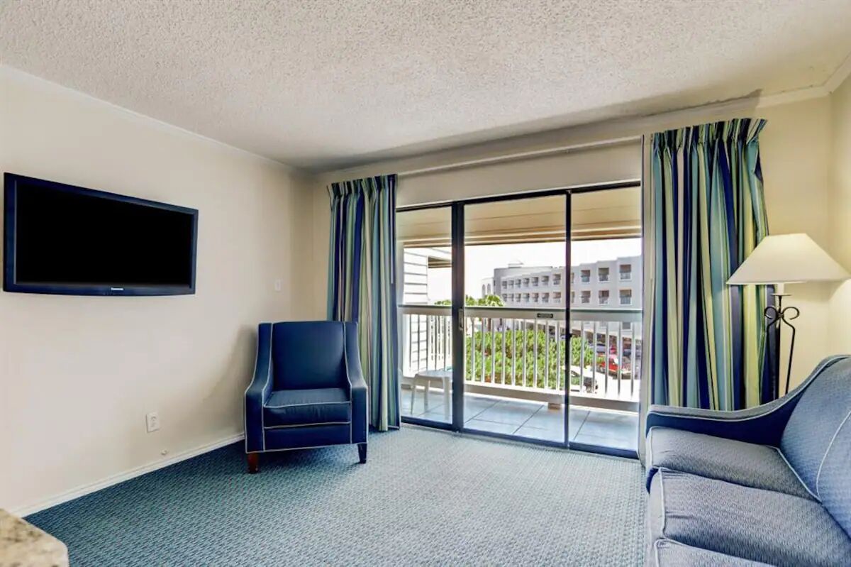 A hotel room with a large window, a flat-screen TV mounted on the wall, a sofa, a chair, and a lamp.