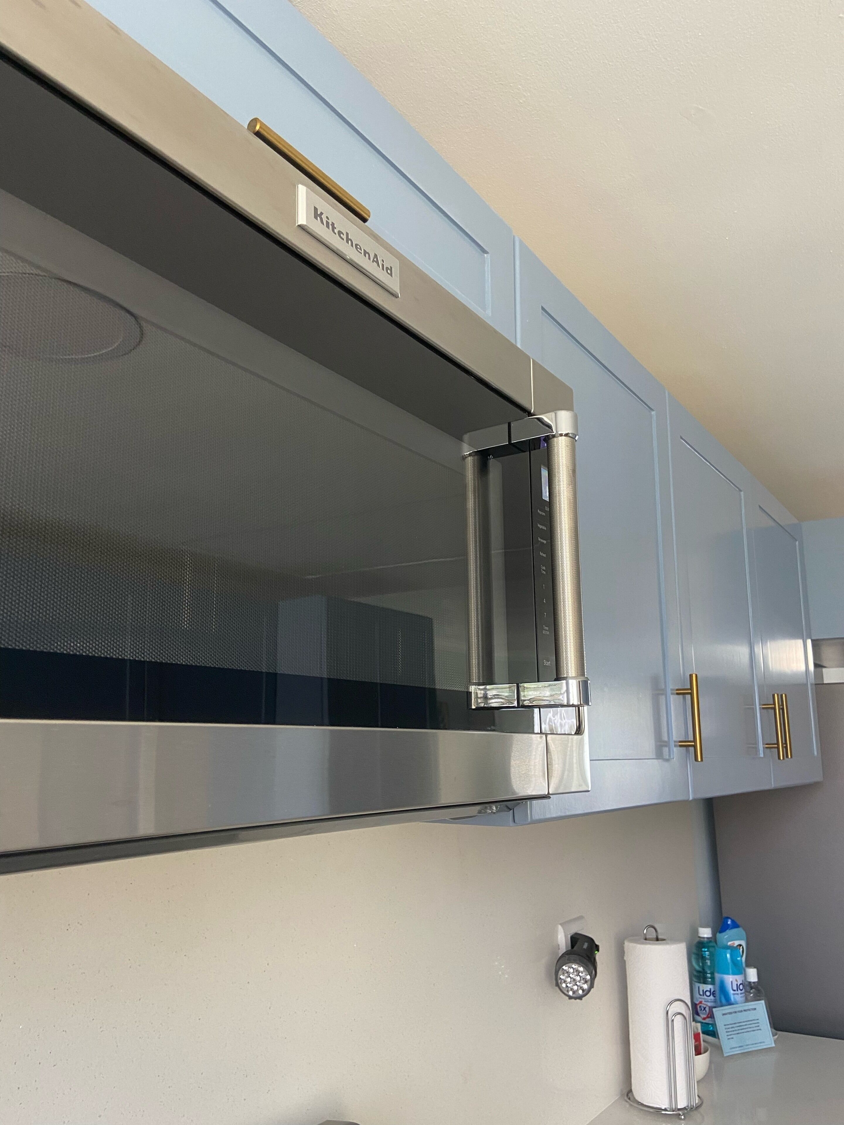Uma cozinha com micro-ondas de aço inoxidável, uma placa com a inscrição 'KitchenAid' e bancada com dispensador de papel toalha, sabonete líquido e um pequeno recipiente.