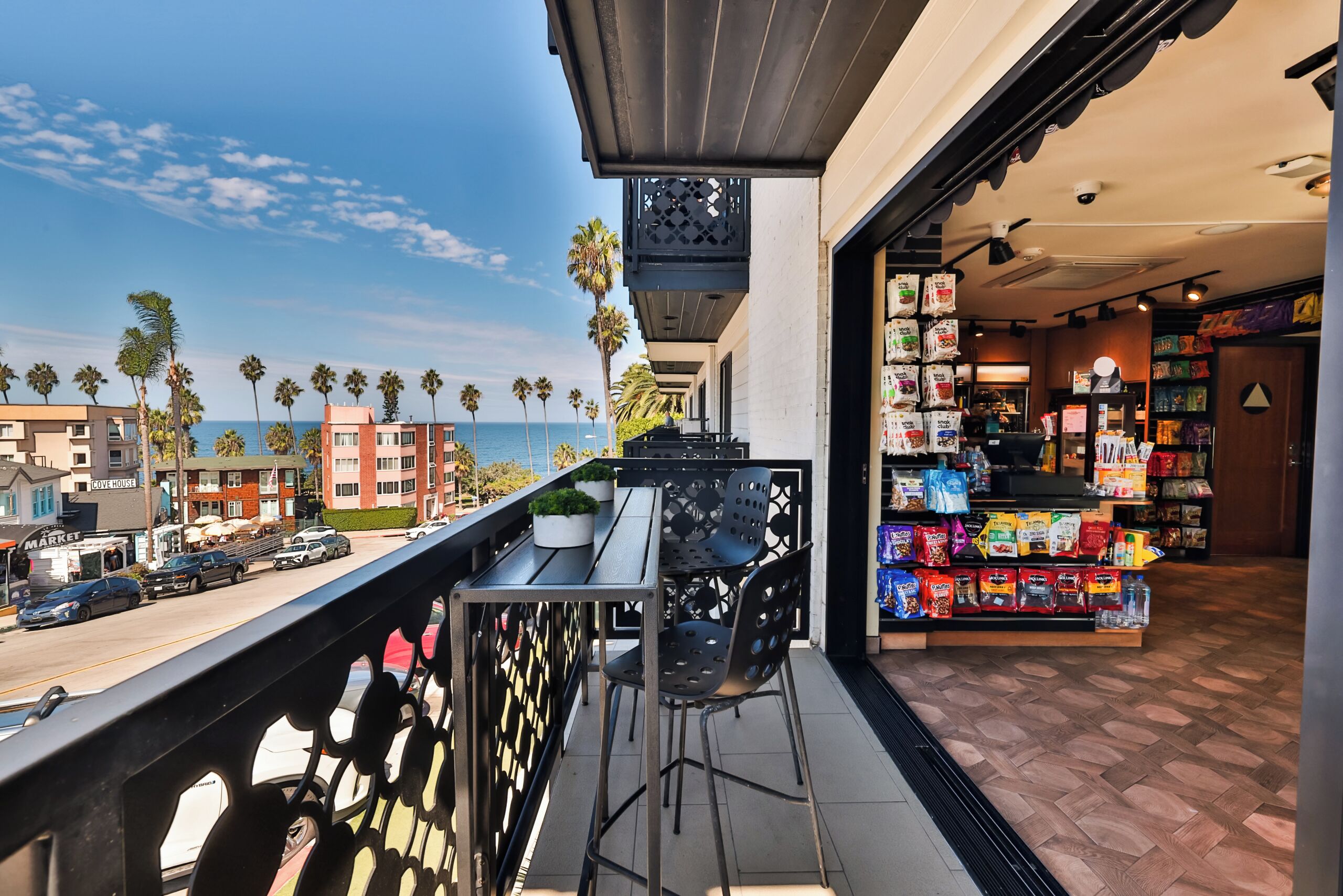 A balcony with a view of the ocean and palm trees, a store with snack bags, and a street with parked cars.