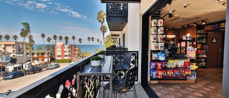 A balcony with a view of the ocean and palm trees, a store with snack bags, and a street with parked cars.