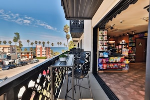 Un balcón con vista al océano y palmeras, una tienda con bolsas de snacks, y una calle con coches estacionados.