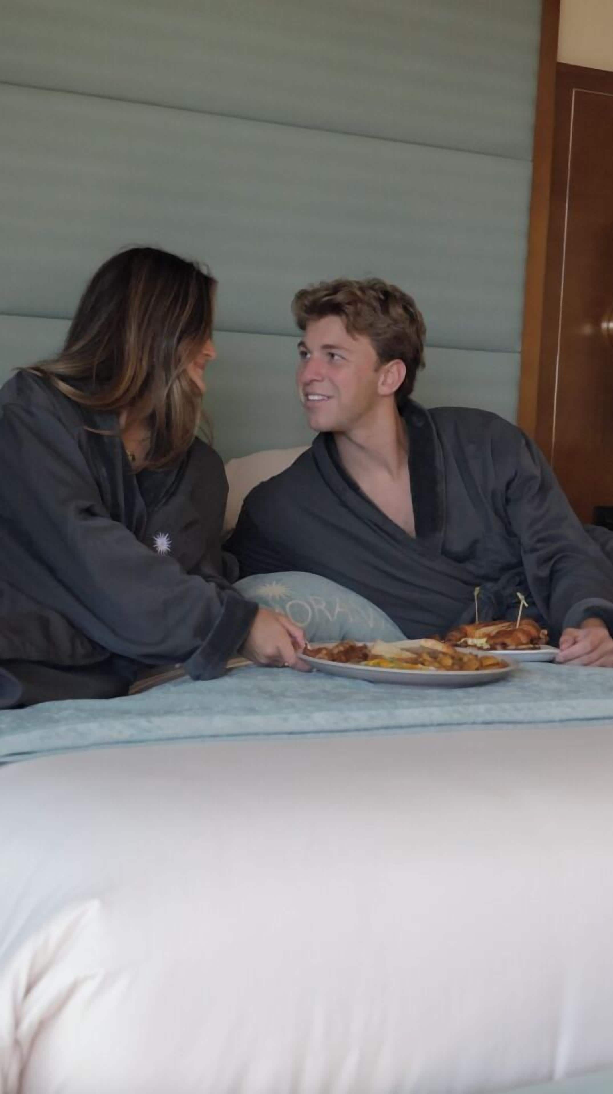 Zwei Personen sitzen auf einem Bett, vor ihnen steht ein Teller mit Essen.