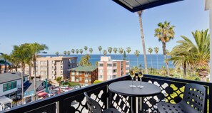 Ein Balkon mit Tisch und Stühlen mit Blick auf einen Strand, im Hintergrund Palmen und Gebäude.