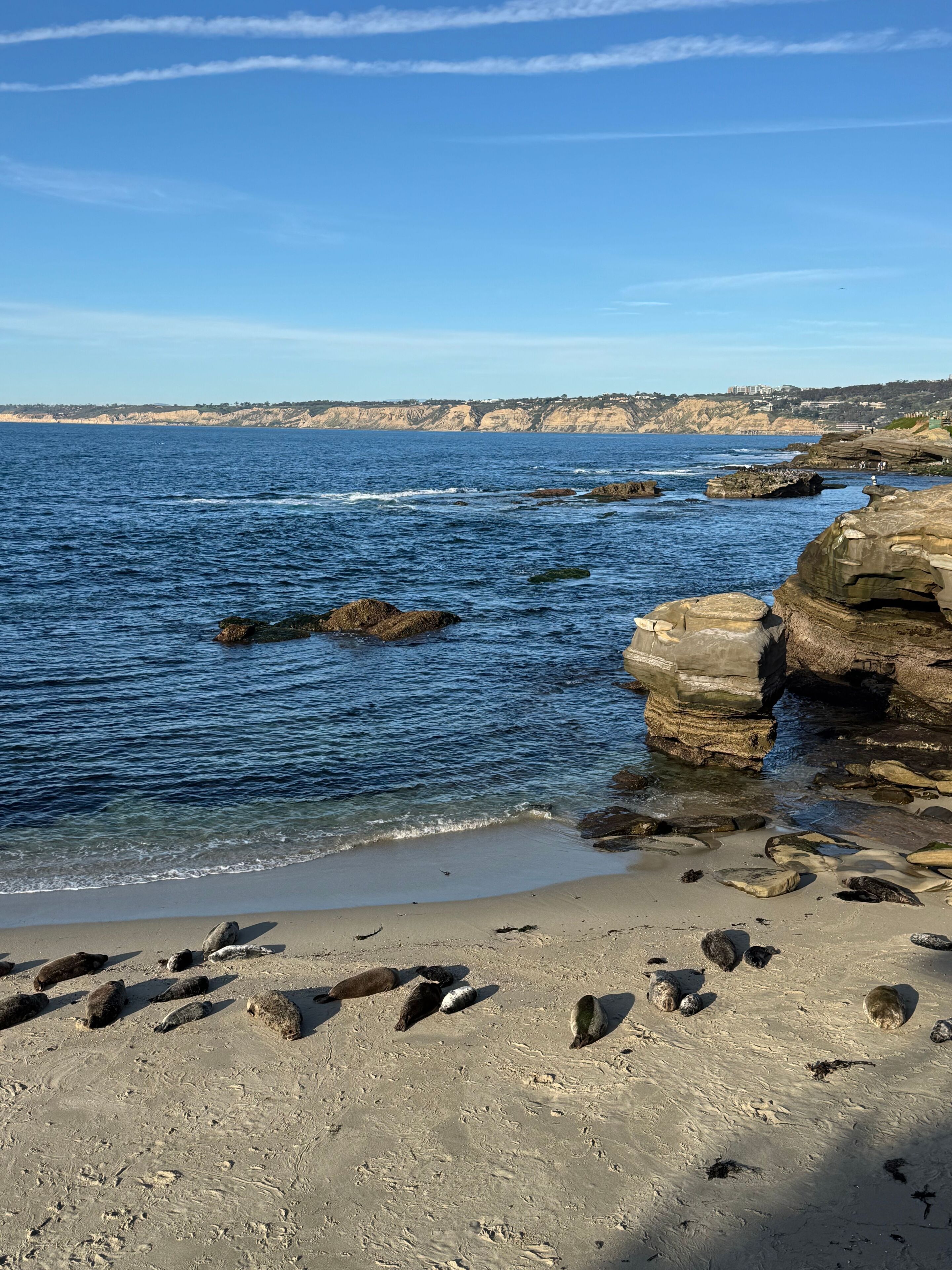 A coastal beach with clear blue water, rocky formations, and a distant cliff with buildings.