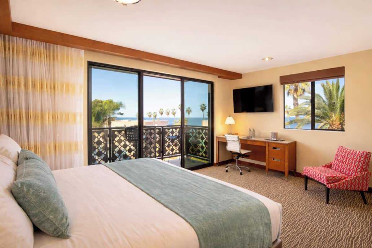 Habitación de hotel con cama, escritorio, silla, televisor y balcón con vista a la playa y palmeras.