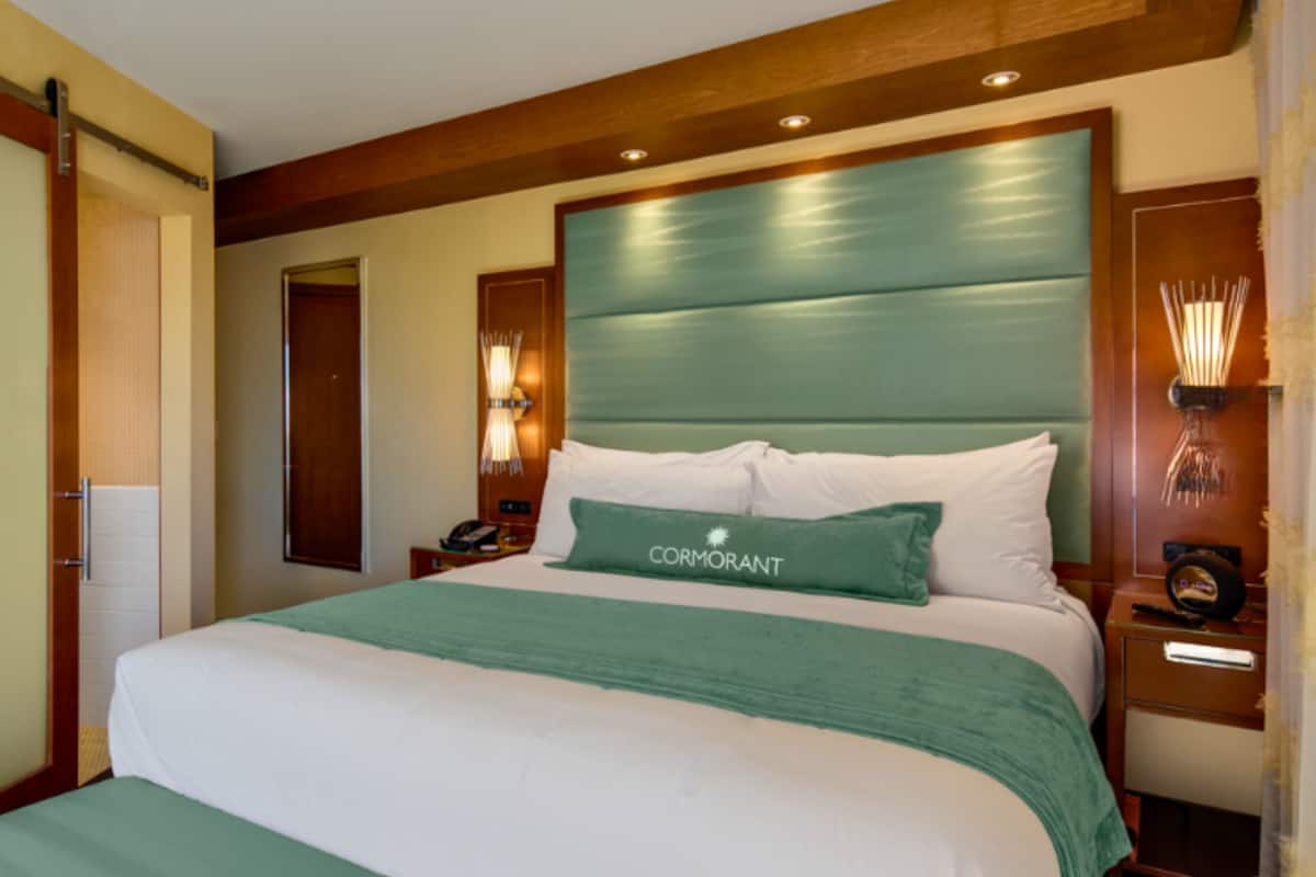 Una habitación de hotel con una cama grande, un cabecero de madera, una mesita de noche con lámpara y una puerta.