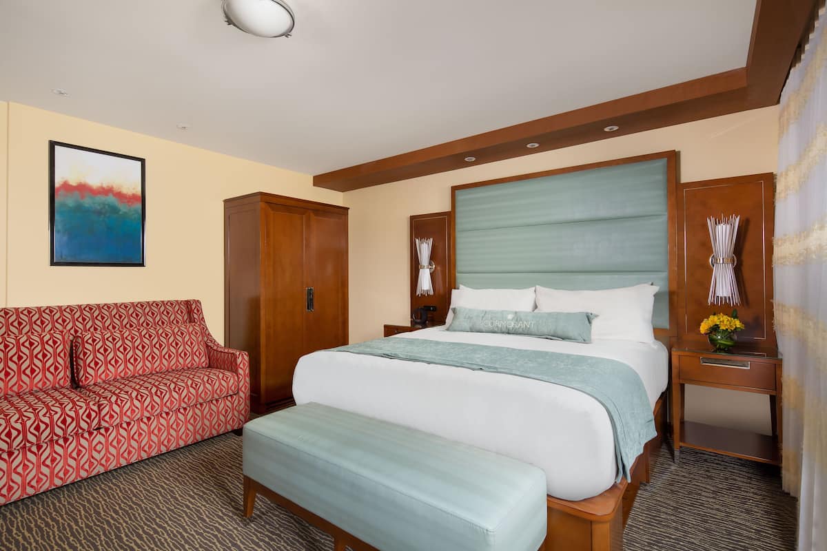 Habitación de hotel con una cama grande, un sofá rojo estampado, un banco de madera, una mesita de noche con un jarrón de flores y un cuadro en la pared.