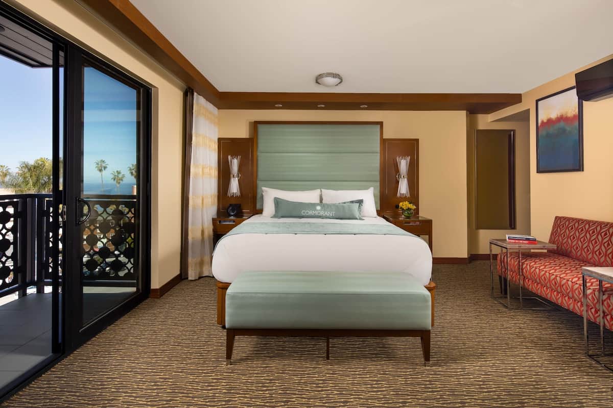 Habitación de hotel con una cama grande, un sofá, una mesita y un balcón con vistas a palmeras.