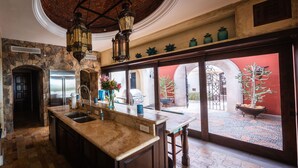A kitchen with a stone wall, a large island with a sink, and a view of a courtyard with a gate and plants. - Long Term Rental Family Style Villa with Oceanview  (San Jose del Cabo)