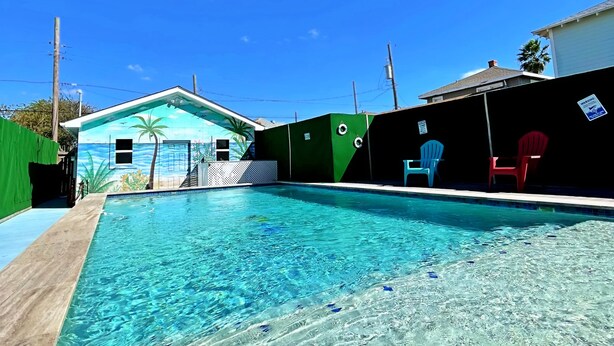 Uma piscina cristalina com um painel tropical ao fundo, cerca verde e uma cadeira vermelha.
