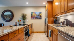 A modern kitchen with granite countertops, wooden cabinets, stainless steel appliances, and a painting of a canyon. - Amazing 2bd/2ba condo!  Custom updates, stellar amenities and prime location! (Fraser)