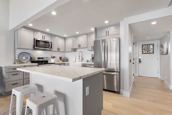 A modern kitchen with white cabinetry, stainless steel appliances, and a marble island with bar stools. - Ma'alaea Surf, D9 - Oceanfront Condo - remodeled two-bedroom, 2nd Floor (Kihei)