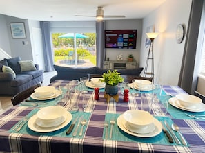 A dining area with a table set for four, a sofa, a TV displaying various channels, and a view of the outdoors.