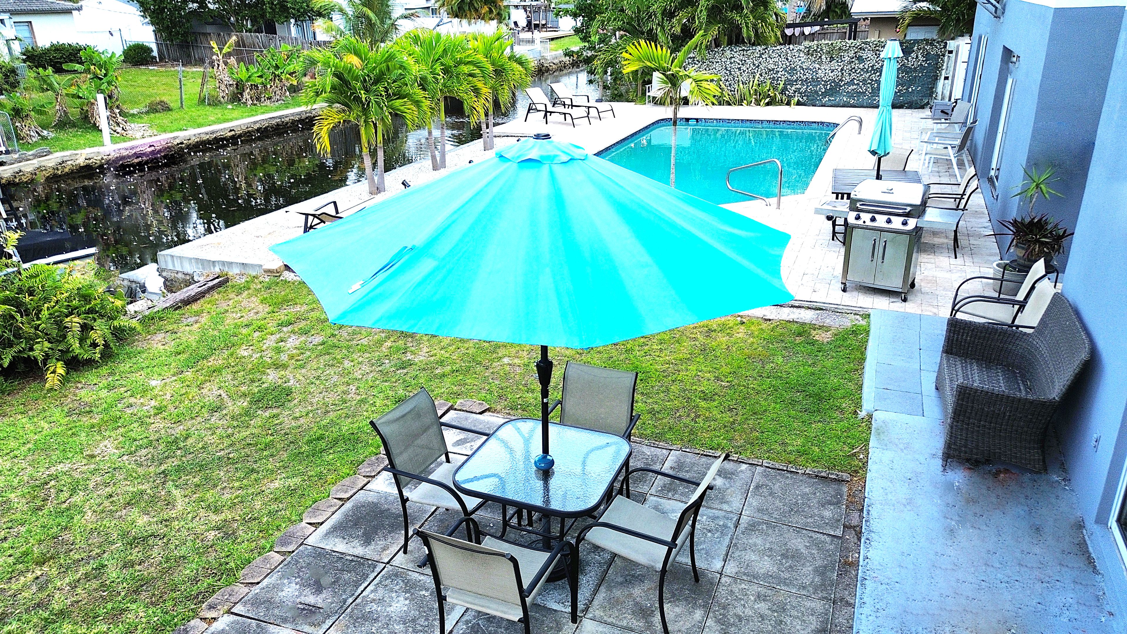 A patio with a table and chairs, a blue umbrella, and a swimming pool.