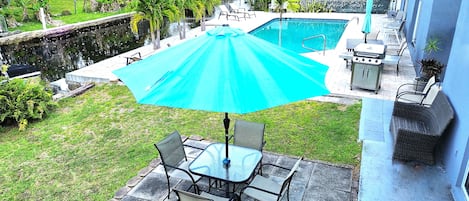 A patio with a table and chairs, a blue umbrella, and a swimming pool.