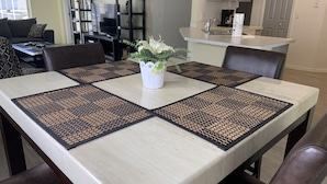 A dining table with four placemats, a vase with flowers, and a kitchen in the background. - Summer State Of Mind - NEW Pool Home (Haines City)