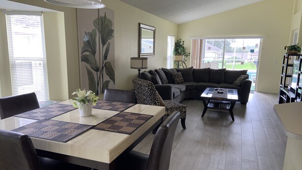 A living room with a dining table, sofa, and coffee table. There is a large window with blinds, a lamp, and a plant. - Summer State Of Mind - NEW Pool Home (Haines City)