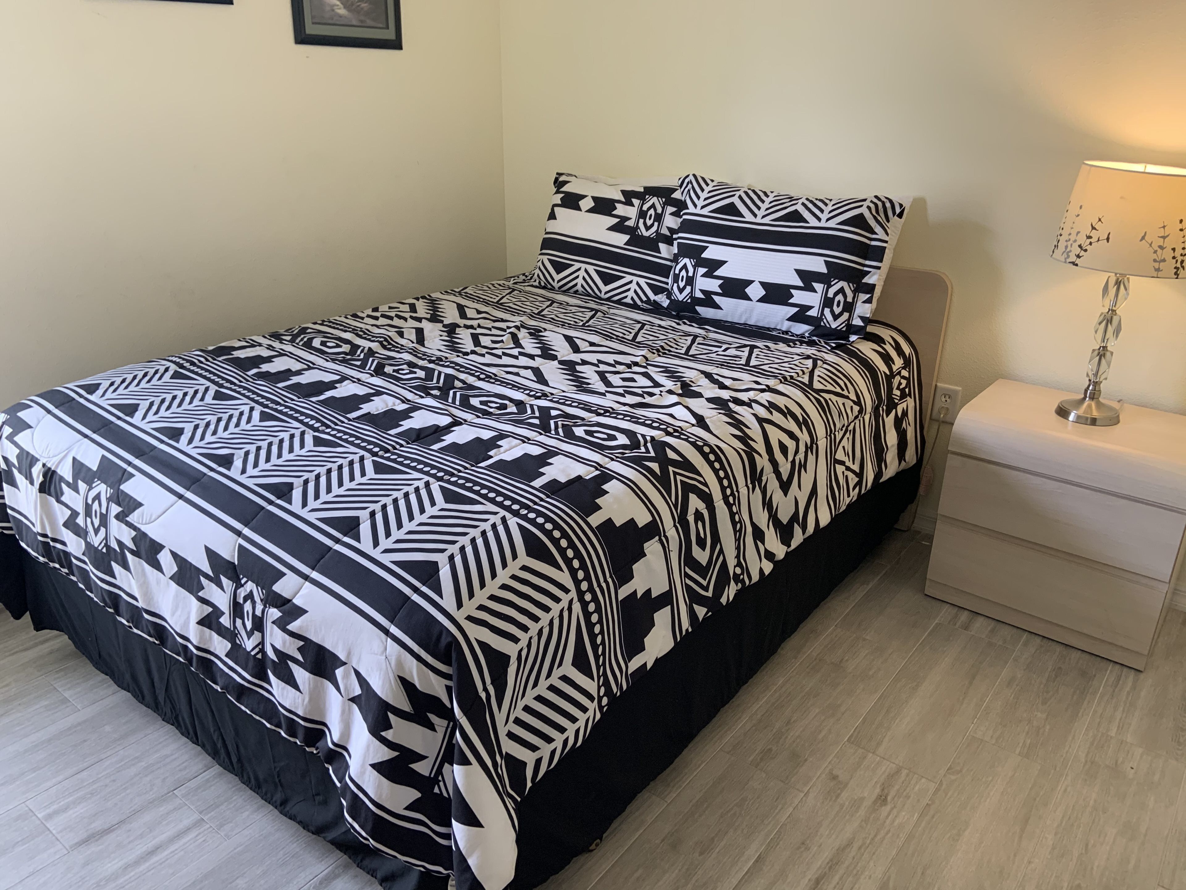 A bed with a black and white patterned bedspread and pillows, a bedside table with a lamp, and a light-colored wall.