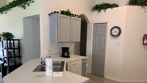 A compact kitchen with a sink, dishwasher, and coffee maker. There is a door with a clock on the wall and a fire extinguisher mounted on the wall. - Summer State Of Mind - NEW Pool Home (Haines City)