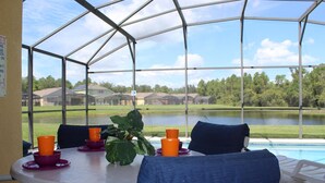 A covered patio with a table and chairs, a view of a lake, and houses in the background. - Just Keep Swimming - NEW Pool Home (Davenport)