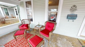 A covered porch with wicker furniture and red cushions, a small table with a bottle, and decorative wall art.