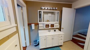 A bathroom with a white vanity, a mirror with lights, a towel rack, and a door leading to a staircase.