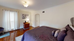 Un dormitorio con cama, cómoda, lámpara, puerta y ventana con cortinas.