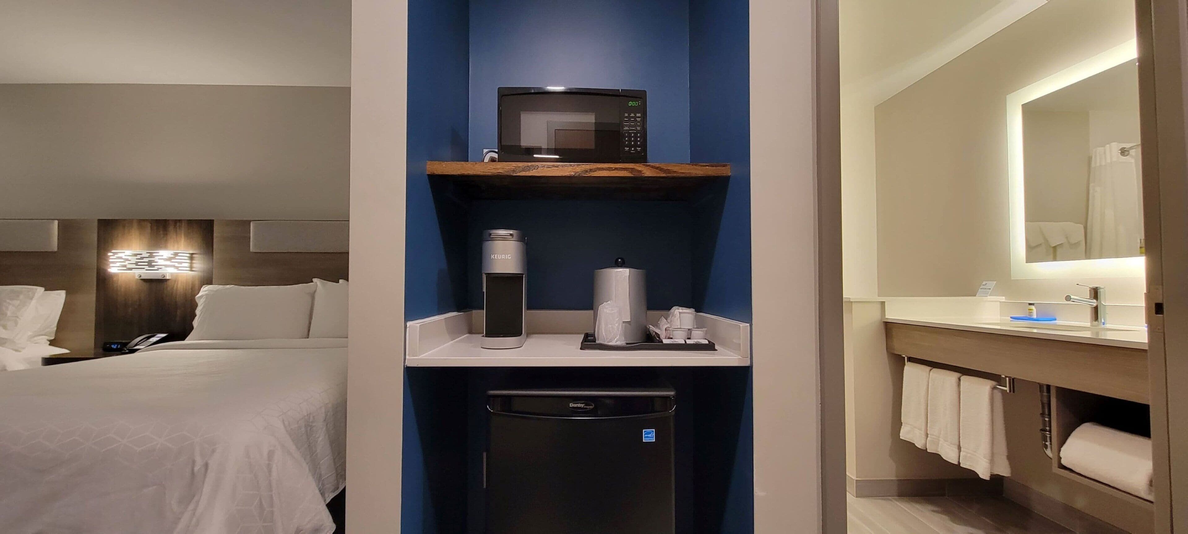 A hotel room with a bed, a minibar, a coffee machine, a trash bin, and a bathroom with a mirror and towel rack.