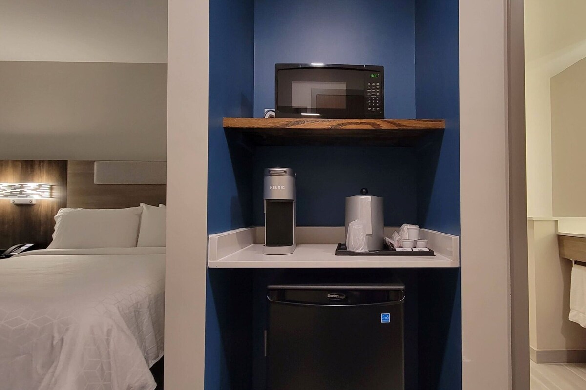 A hotel room with a bed, a minibar, a coffee machine, a trash bin, and a bathroom with a mirror and towel rack.