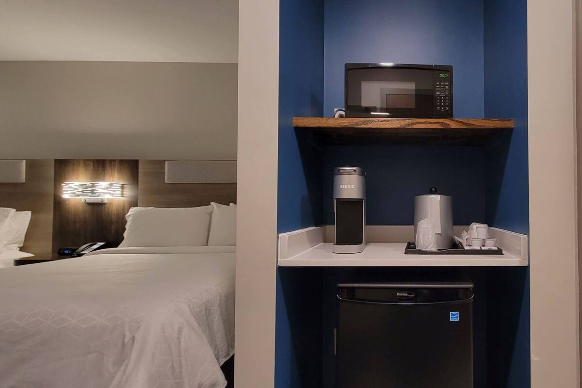 A hotel room with a bed, a minibar, a coffee machine, a trash bin, and a bathroom with a mirror and towel rack.