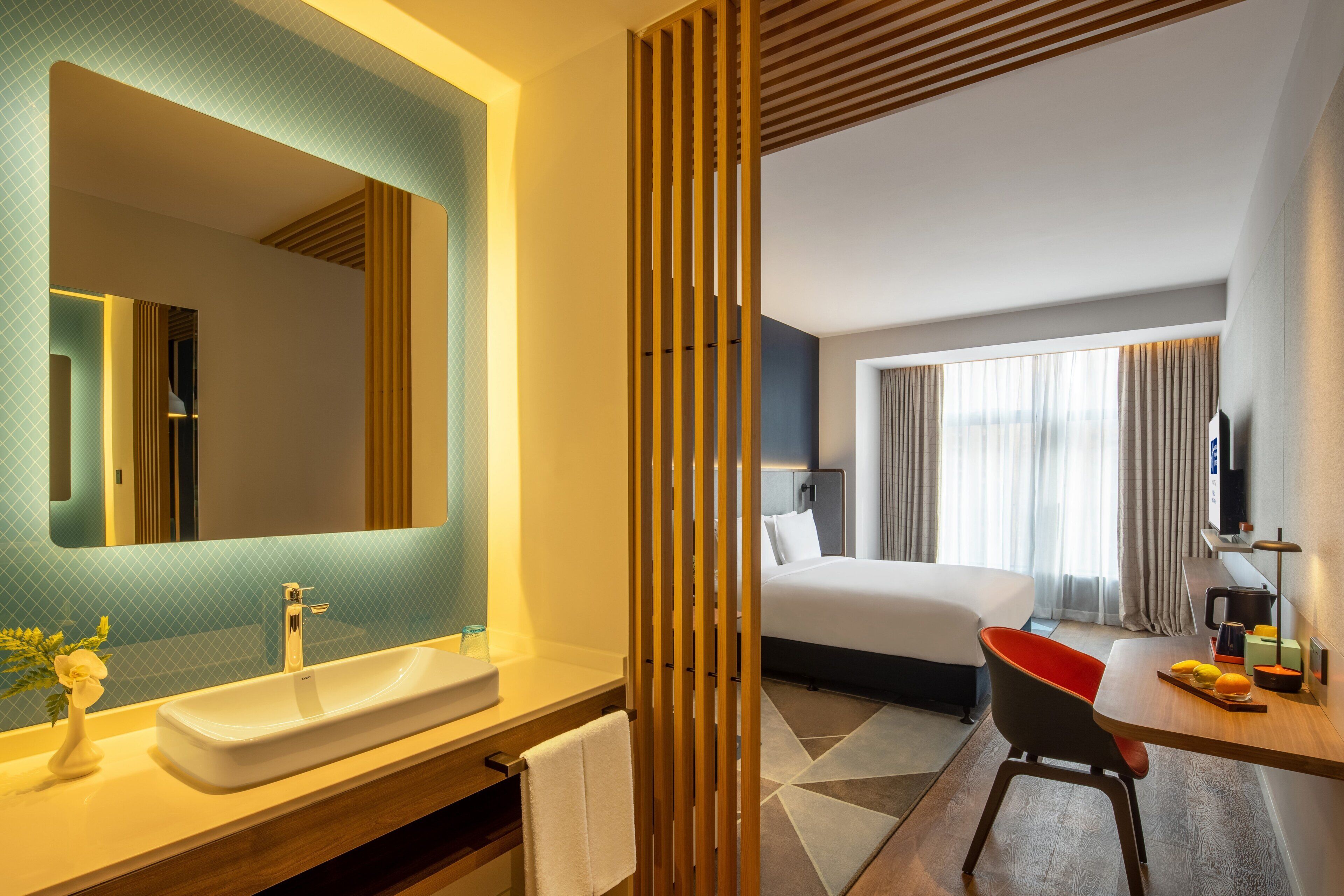 Quarto de hotel com um espelho grande, pia, cama, escrivaninha e cadeira, e uma televisão.
